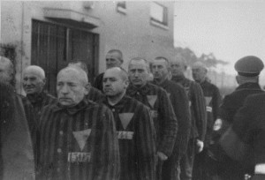 Ένστολοι κρατούμενοι, που φέρουν στα ρούχα τους τριγωνικά σήματα, συναθροίζονται υπό τη συνοδεία Ναζί φρουρών στο στρατόπεδο συγκέντρωσης Σαξενχάουζεν. Σαξενχάουζεν, Γερμανία, 1938. — National Archives and Records Administration, College Park, Md. Πηγή: https://www.ushmm.org/outreach/el/media_ph.php?ModuleId=10007720&MediaId=893 